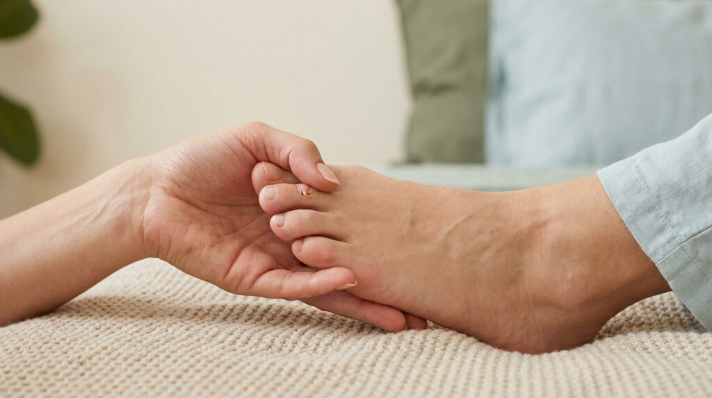 Gros plan d'une main manipulant délicatement l'orteil tordu d'un pied posé sur une couverture beige. Scène de soin ou d'examen podologique.